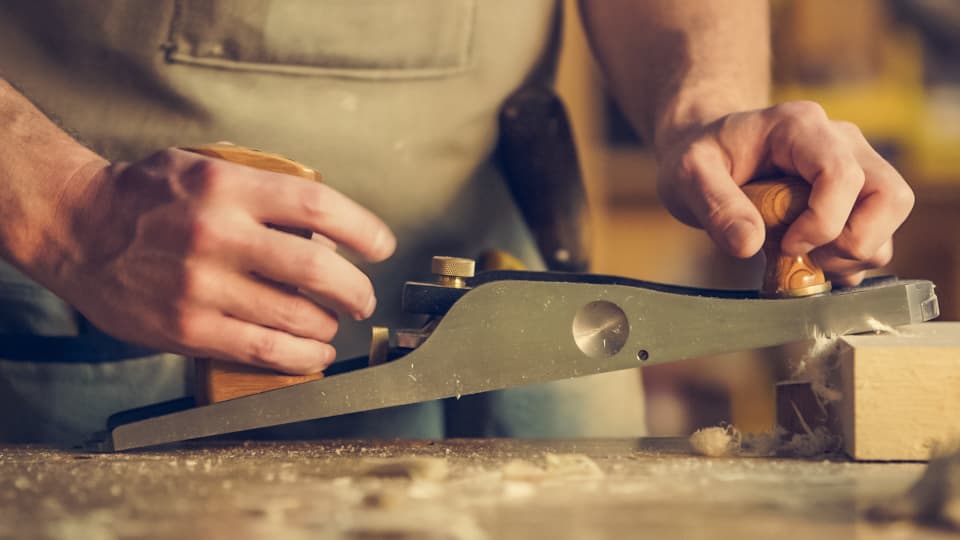 A craftsman's hands running a hand plane along a length of timber, shavings curling off the blade.
